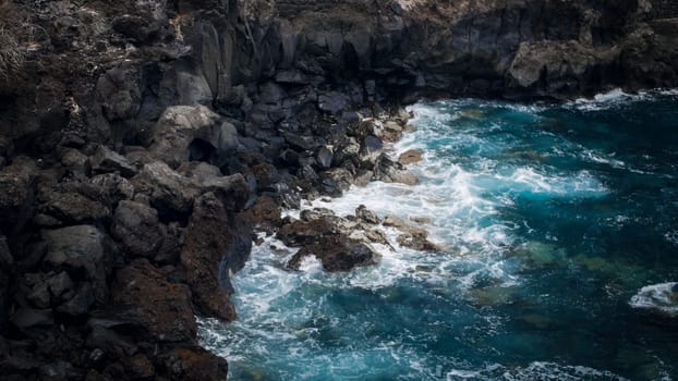 Seascape of blue ocean waves rolling over black volcanic rocks and cliffs on the shore.