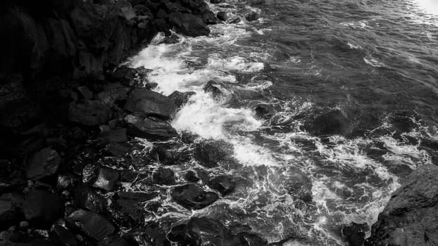 Black and white image of ocean waves rolling and breaking on sharp cliffs and rocks on the beach