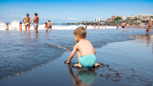 Little toddler boy sitting on the beach and playing with ocean waves.