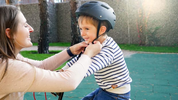 Portrait of smiling young mother adjusting and fastening her son protective helmet buckles before riding bicycle.