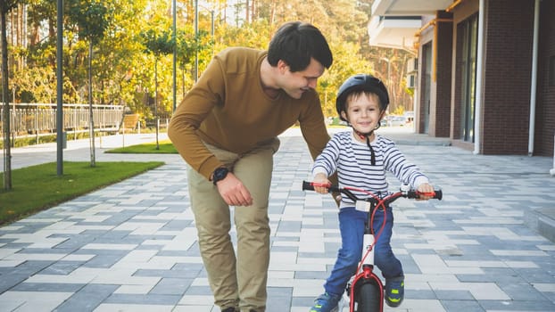Toned image of happy smiling little boy riding his first bicycle with father walking near him.