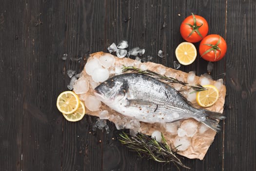 Raw dorada fish or gilt-head bream on ice with lemon slices and rosemary over black wooden background, flat lay, top view