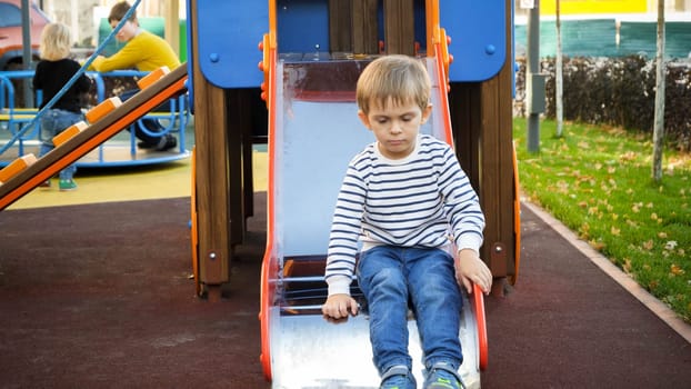 Sad and upset little lonely boy sitting on slide at playground.
