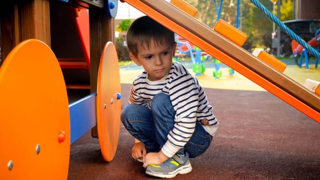 Sad little boy hiding under slide on children playground at kindergarten.