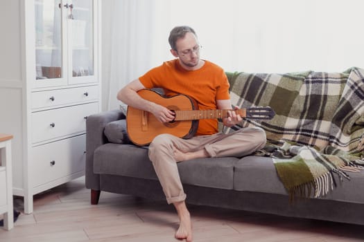 Young caucasian man playing the guitar sitting on couch at home. Practice music, lifestyle photo.