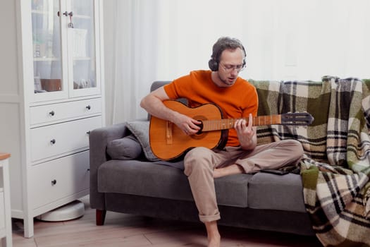 Young caucasian man in headphones playing the guitar sitting on couch at home. Practice music, lifestyle photo