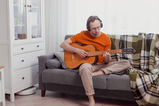 Young caucasian man in headphones playing the guitar sitting on couch at home. Practice music, lifestyle photo