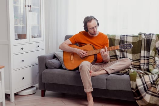 Young caucasian man in headphones playing the guitar sitting on couch at home. Practice music, lifestyle photo