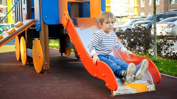Upset little boy sitting on the slide at children playground.