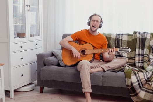 Young caucasian man in headphones playing the guitar and singing at home. Quarantine and hobbies concept. Practice music, lifestyle photo