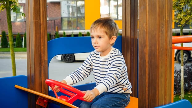 Happy smiling boy playing on children playground at park and turning steering wheel of wooden car.