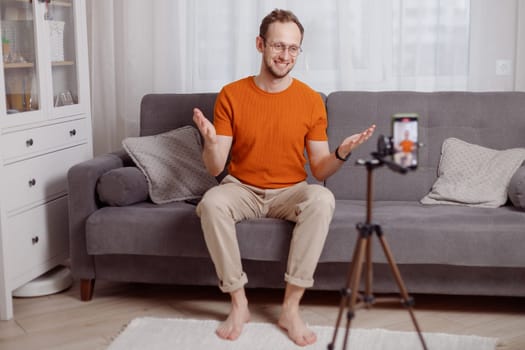 Hipster man blogger recording vlog on mobile phone sit on couch in living room and explain and gesturing with hands.