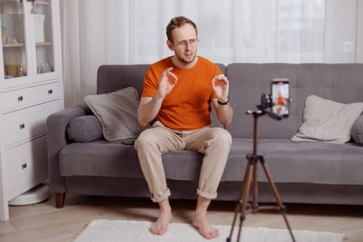 Hipster man blogger recording vlog on mobile phone sit on couch in living room and explain and gesturing with hands.
