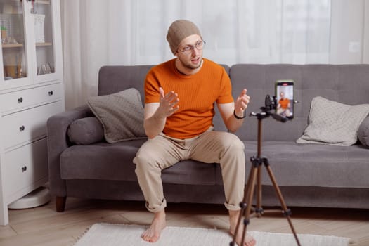 Hipster man blogger recording vlog on mobile phone sit on couch in living room and explain and gesturing with hands.