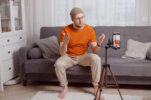 Millennial hipster man blogger recording vlog on mobile phone sit on sofa in living room and explain and gesturing with hands. Doing content for social media.