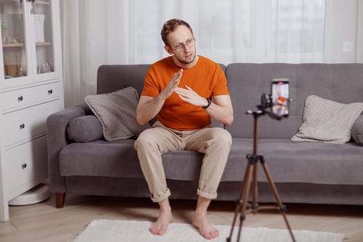 Hipster man blogger recording vlog on mobile phone sit on couch in living room and explain and gesturing with hands.