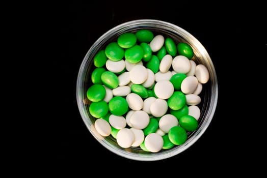 A round glass bowl filled with green and white mints of circular shape on a black isolated background.