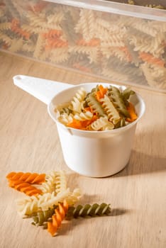 Three different colours of fussily fill a white plastic measuring cup, with a plastic container filled with more fussily in the back and some pieces of fussily lying on the wooden surface right in front.