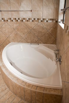 A white corner bath in a tiled bathroom, the tiles are of different shades of brown