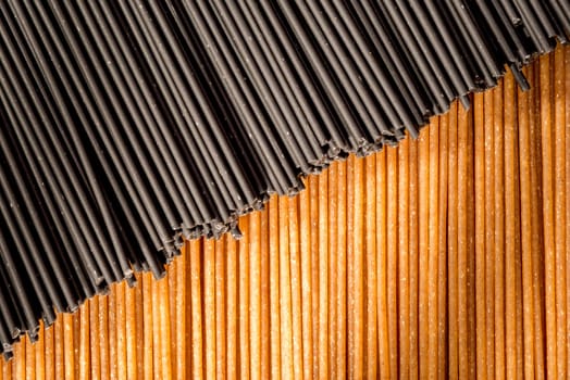Two different types of raw spaghetti with the black spaghetti places diagonally across the brown spaghetti to form an abstract background image with natural window light falling on the pasta.