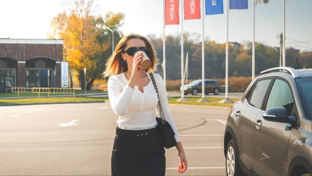 Portrait of young businesswoman walking on car parking and drinking coffee from paper cup.
