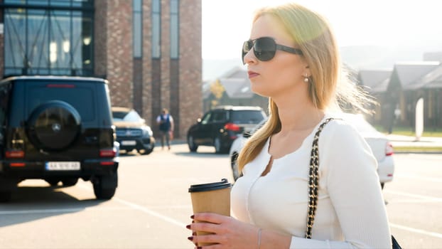 Portrait of beautiful blonde girl drinking coffee and walking on car parking.