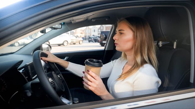 Portrait of beautiful blonde woman driving a car and drinking coffe from paper cup.