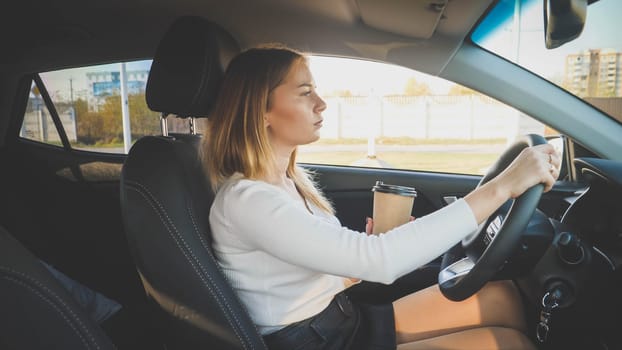 Toned portrait of elegant young woman driving a car and holding coffee in paper cup.