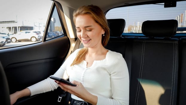 Portrait of smiling young businesswoman typing messafe on smartphone while riding to work in taxi car.