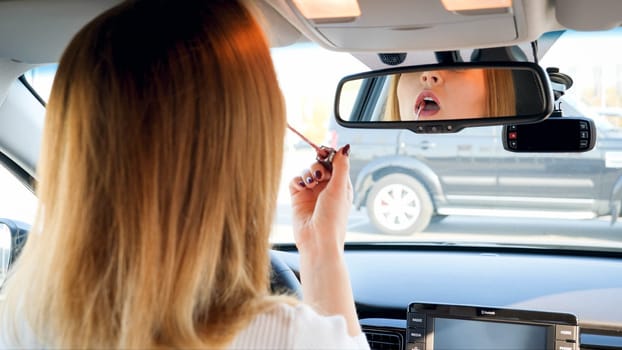 Portrait of beautiful young woman looking in car mirror while painting her lips with lipstick.