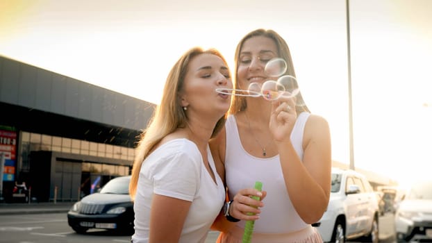 Two young women having fun and blowing soap bubble on city street.