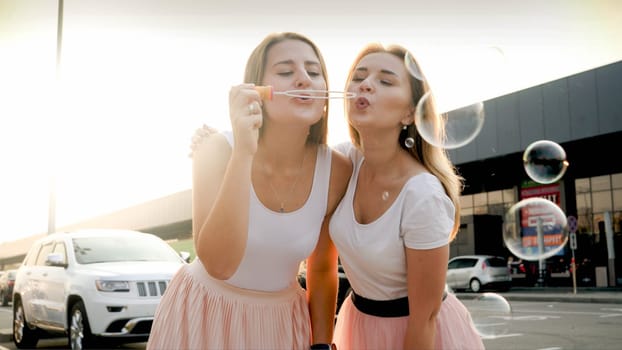 Portrait of two beautiful girls blowing soap bubbles and having fun on city street at sunset.