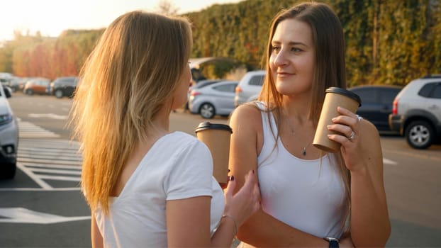 Portrait of two beautiful girls drinking coffee from paper cars while talking on car parking at shopping mall.