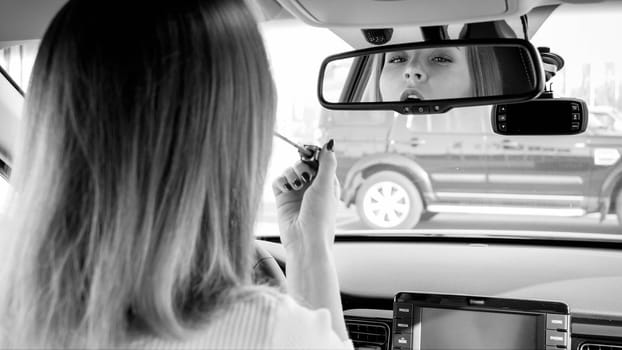 Black and white portrait of beautiful young woman applying makeup while driving a car.
