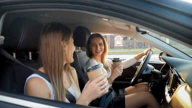 Two smiling girls with coffee paper cups riding in car at sunset.