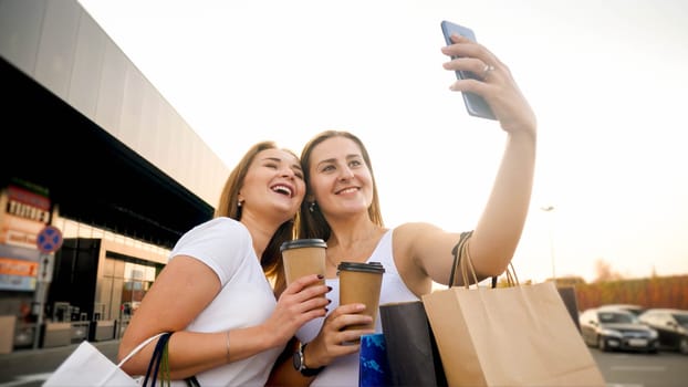 Portrait of two smiling girls making selfie with coffee cups and paper bags at big shopping mall.