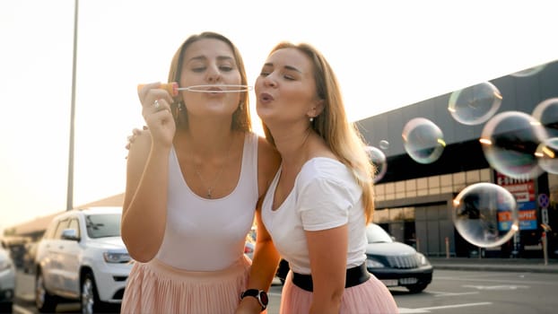 Two happy smiling girl friends blowing soap bubbles on shopping mall car parking.