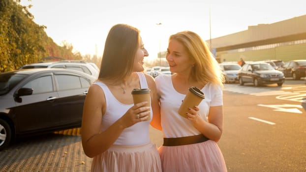 Portrait of two smiling young girls drinking coffee-to-go fro paper cups on shopping mall car parking at sunset.
