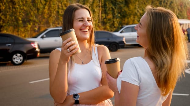 Portrait of two girls drinking coffee, talking and laughing on car parking.