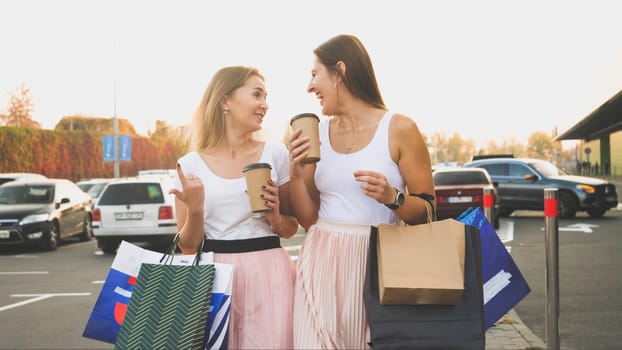 Toned photo of two women with paper bags talking and smiling while walking on car parking at shopping mall.
