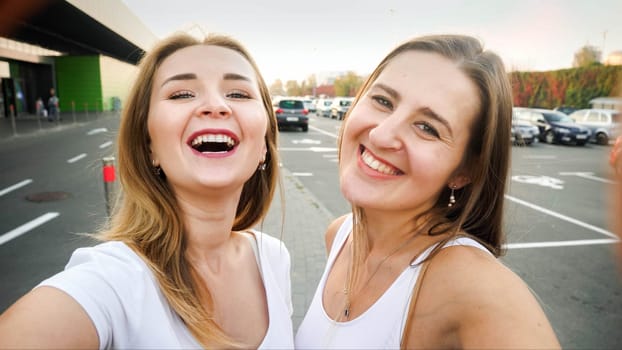 Closeup portrait of two happy smiling young women making selfie on car parking at shopping mall entrance.
