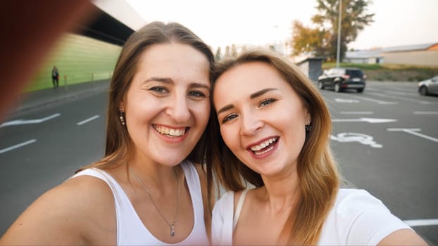 Closeup photo of brunette and blonde smiling girls making selfie on smartphne at car parking.