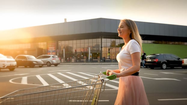 Beautiful smiling blonde girl pushing shopping cart while walking on car parking at shopping mall.