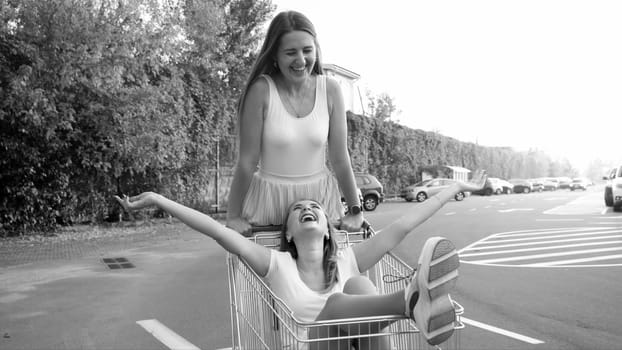 Black and photo of two laughing girls friends riding in shopping trolley on car parking at shopping mall.