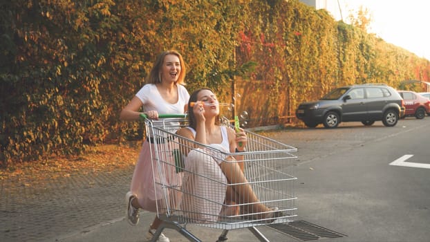 Toned photo of two happy girls riding in shopping cart and blowing soap bubbles on car parking.