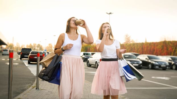 Two beautiful young girls walking with shopping bags and drinking coffee from paper cups.