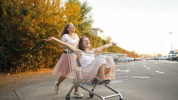 Portrait of two laughing and smiling girls riding and pushing each other in shopping cart on shopping mall car parking.