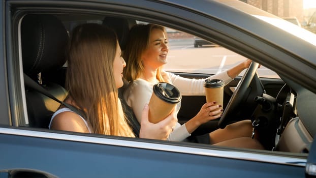 Portrait of two smiling girls driving in car and drinking coffee from paper cups at early morning.