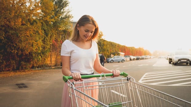 Toned portrait of beautiful smiling young girl walking with shopping trolley on car parking.
