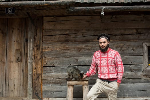 portrait of young hipster man with beard and cat in front of wooden house